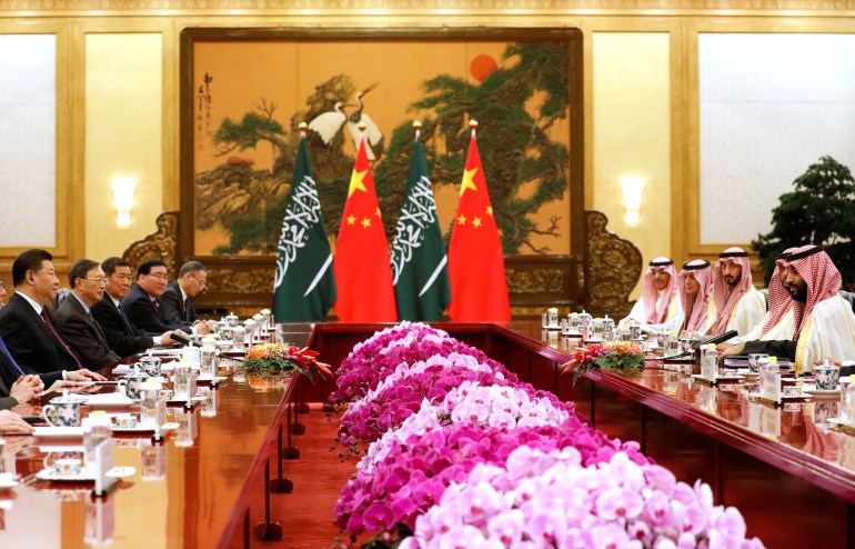 Saudi Crown Prince Mohammad Bin Salman faces Chinese President Xi Jinping across a bank of light and dark pink flowers during a summit in Beijing. Xi and his entourage are sitting along a table on one side and MBS and his entourage on the other. There is a traditional Chinese painting at the back and the flags of Saudi Arabia and China