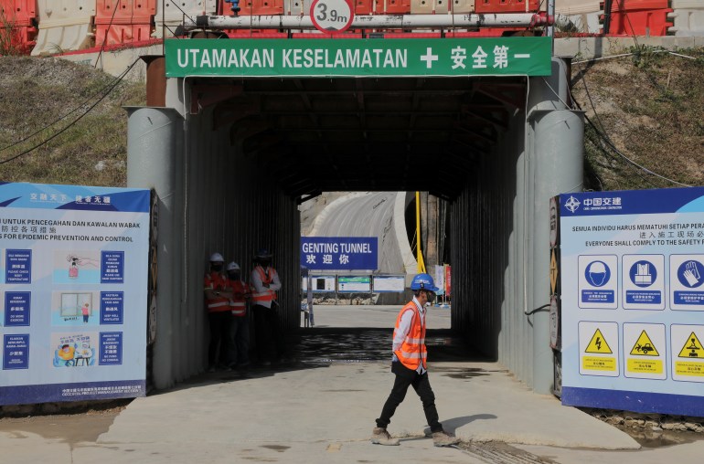 A worker in a high-vis jacket and hard hat walks past a tunnel entrance at the construction site of the ECRL in Malaysia. There are signs saying 'safety first' in Malay and Chinese.