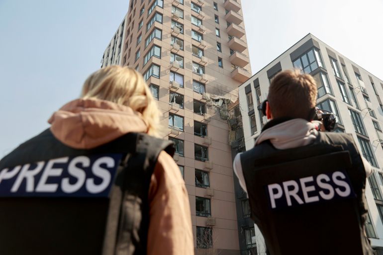 Journalists look up at damaged high-rise building, both wear bullet proof vests that say "PRESS" on the back