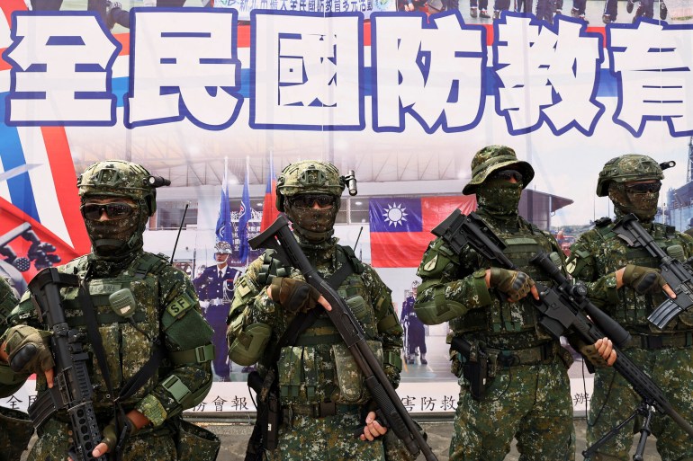 Quatre soldats faisant la queue pour une photo en uniforme de combat complet lors des exercices de défense civile « Minan » à Taïwan.  Il y a une bannière montrant des drapeaux de Taïwan avec de grands caractères chinois derrière eux