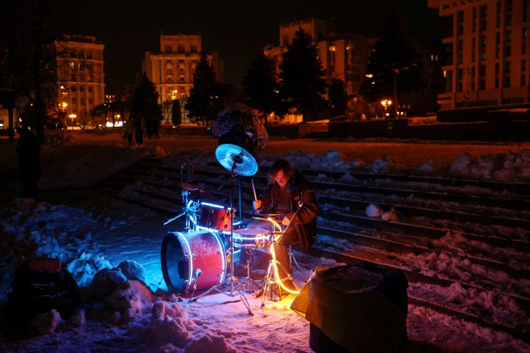 Um músico senta-se na neve em Kyiv com sua bateria iluminada por luzes e os prédios ao redor na escuridão.