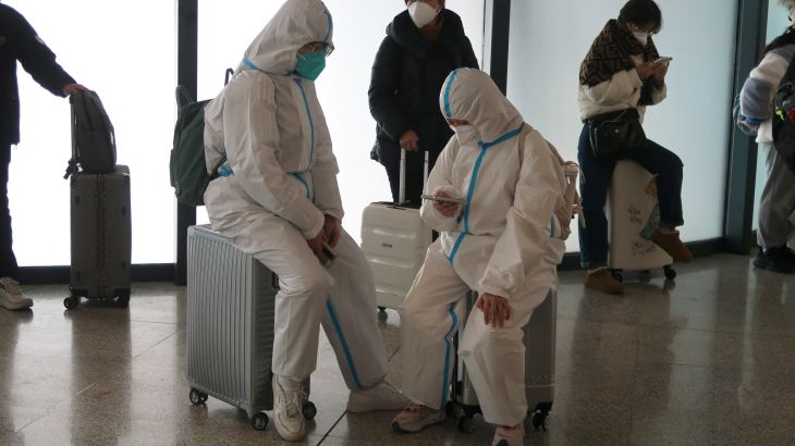 Travellers in full white hazmat suits sit on their luggage as they wait to board a train. One is looking at their phone