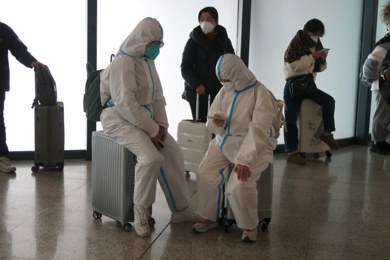 Travelers in all-white hazmat suits sit on their luggage as they wait to board the train.  A person is looking at their phone.