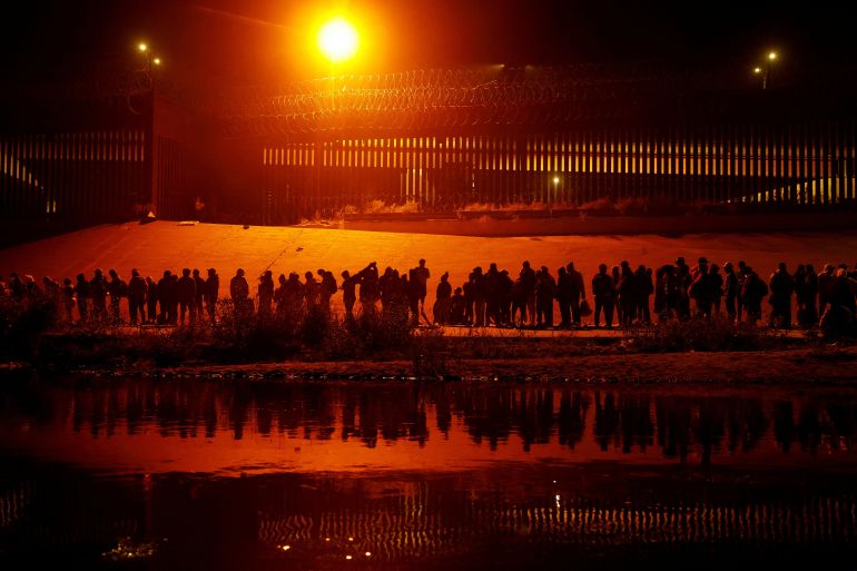 Refugees and migrants, silhouetted against a bright orange sun, line up along the Rio Grande River.