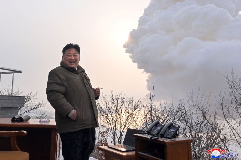 N Korean leader Kim Jong Un watches the test of a 'high thrust solid fuel motor'. He's wearing a thick brown winter jacket and appears to be standing on a hill. He looks very pleased and is smiling. There are large clouds of smoke