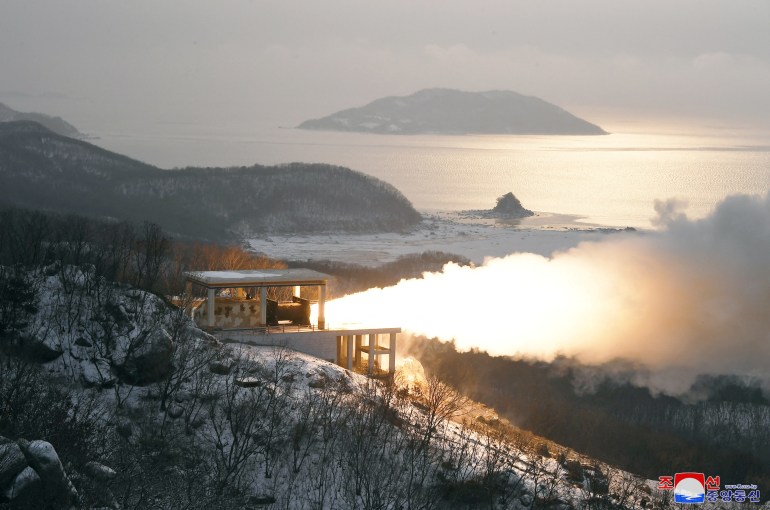 A view of a "high-thrust solid-fuel motor" test. Smoke and flames are shooting out of a a device on a hillside. There is a small island in the background and the sea. It looks cold