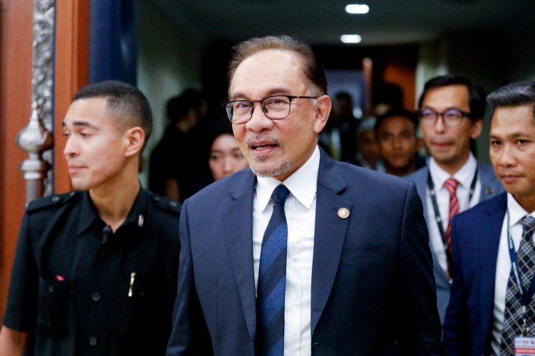 Perdana Menteri Malaysia, Anwar Ibrahim, dengan jas dan dasi biru, berjalan keluar dari koridor dengan orang-orang di belakangnya dan di sampingnya dengan lanyard. 