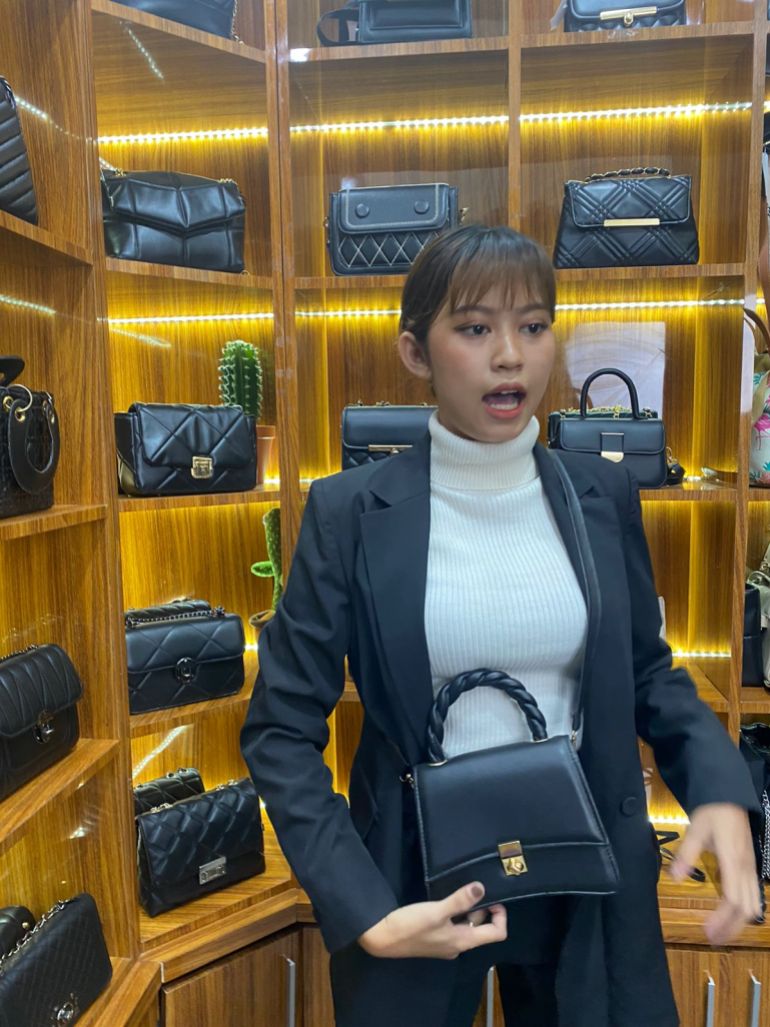 Woman holding hand bag while wearing a business suit with other bags on display on shelves behind her