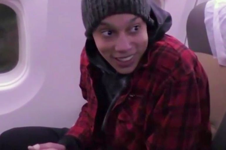 Brittney Griner smiling as she sits on a plane on her way out of Russia. She's wearing a red jacket and grey beanie