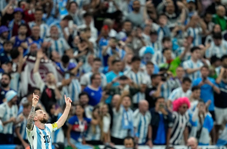 Messi raising his arms in the air and pointing to the sky while looking up against a backdrop of Argentina fans wearing the white and blue stripes in the stands