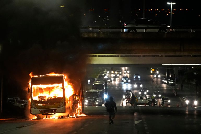 A bus engulfed in flames on the roadside in front of a flyover. It's dark and people are silhouetted in the lights and flames.