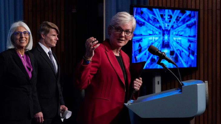 Secretary of Energy Jennifer Granholm, in a red suit announces the fusion breakthrough. There is a screen behind her.