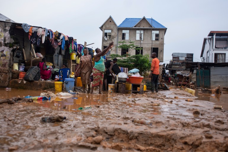 A woman stands in the mud outside her home following floods in Kinshasa. She has her arms up and looks like she's explaining something to a man standing nearby and a woman beside her. There is a large pool of muddy water, buckets, a suitcase and other items. Behind is a small tree and a large house with a blue roof