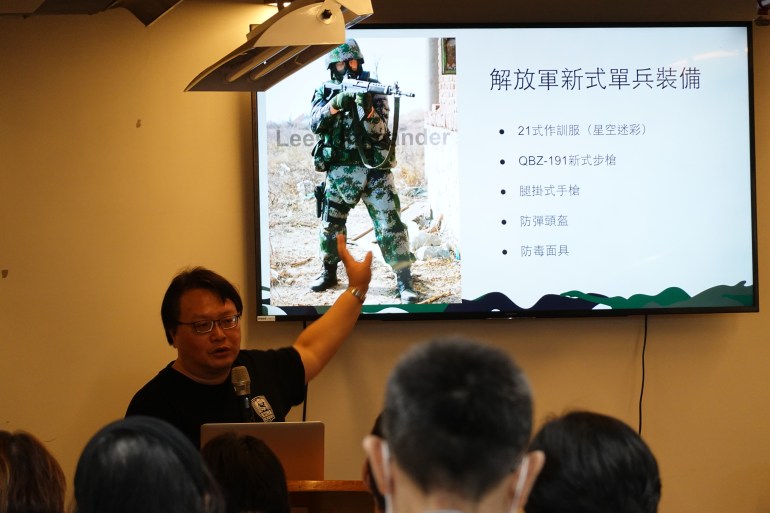 Cheng Hui-Ho fait une présentation aux membres du public sur l'équipement porté par les soldats de l'APL.  Il y a une diapositive sur le mur montrant un soldat avec son arme levée et écrivant en caractères chinois sur le côté droit.  Cheng pointe le toboggan, qui est derrière lui