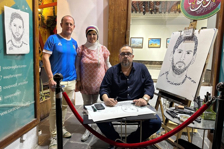 Khaled Almesawy est assis avec un croquis posé sur ses genoux.  Il y a un croquis de Messi sur un chevalet à sa gauche et un autre accroché au mur à sa droite.  Deux personnes posent pour la photo derrière lui.