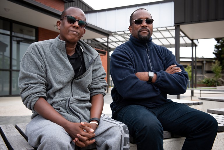 Mahmood from Somalia (left) and Jacques from Cameroon (right), sitting on a bench outside the Mangere Refugee Resettlement Centre in Auckland. They are wearing fleeces and dark glasses and look pensive. Jacques has his arms folded
