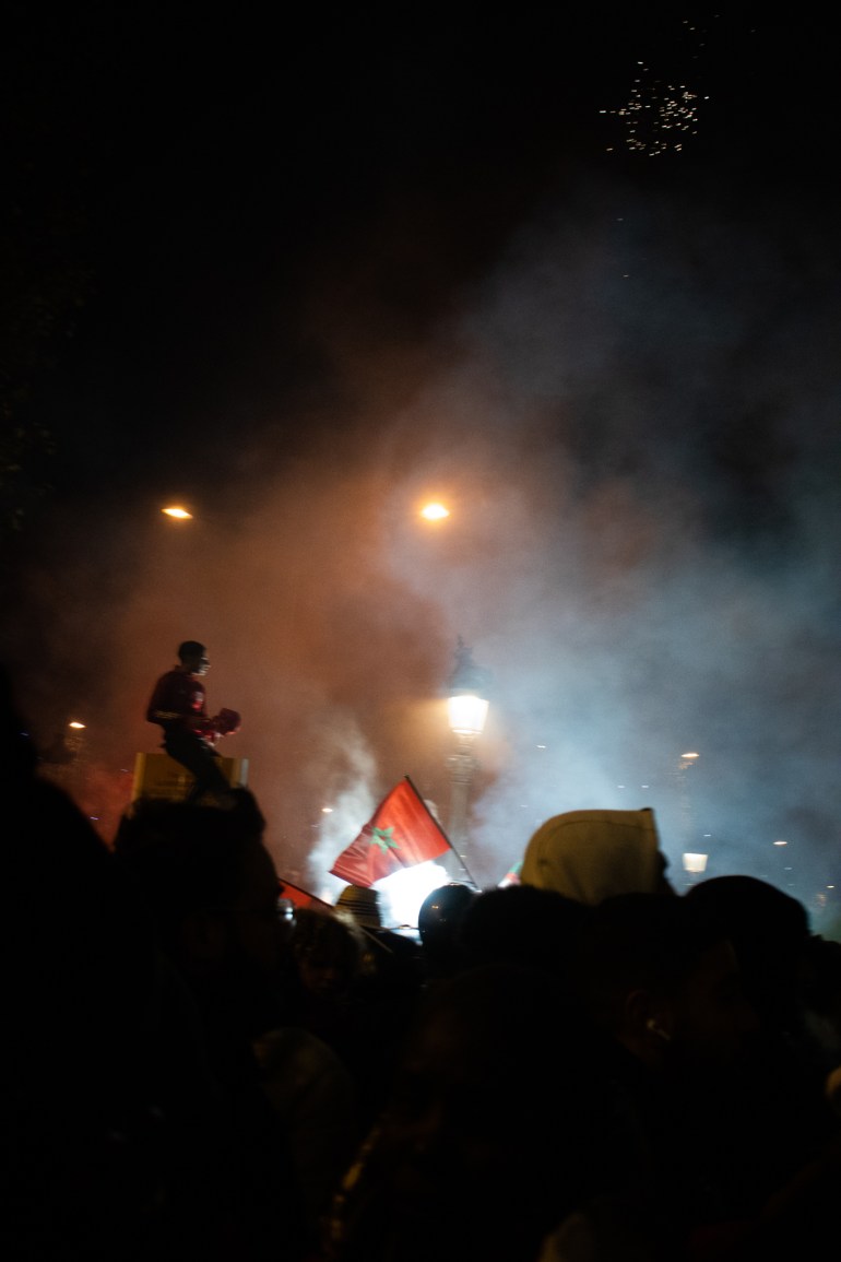 Silhouette of a person sitting while a Moroccan flag is lit in a crowd scene among tear gas
