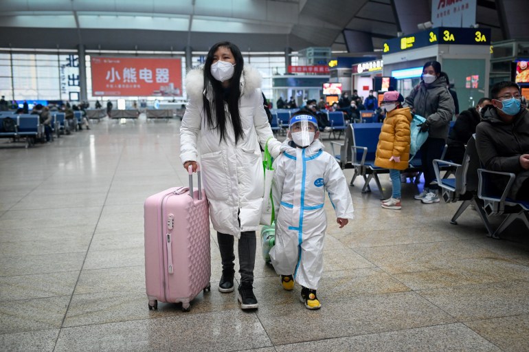 Woman wearing a long white coat, mask and pulling a pink suitcase.  The child next to him is dressed in PPE, a face shield that is visible and wearing a mask.  Other travelers are sitting on side chairs, all wearing masks and coats or warm jackets.