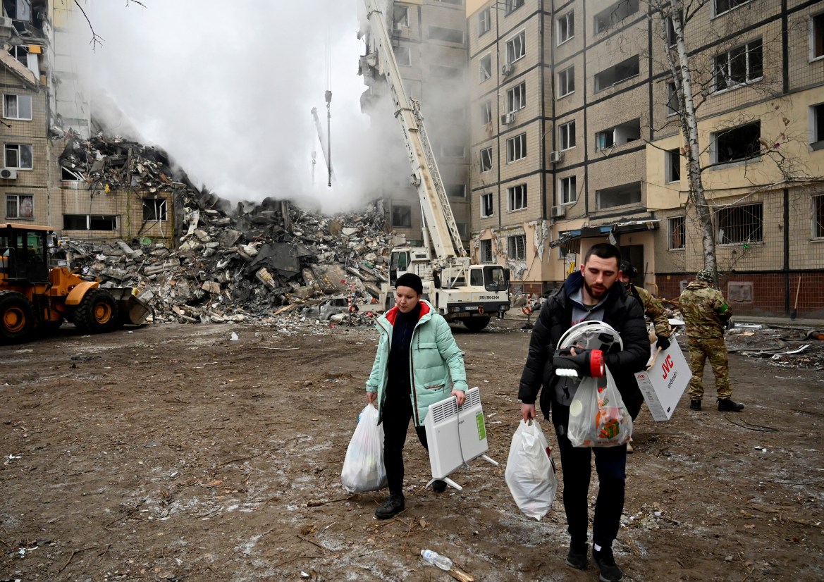 Two residents walk away from the smouldering ruins of a partially destroyed apartment building, with white plastic bags of belongings in their hands.