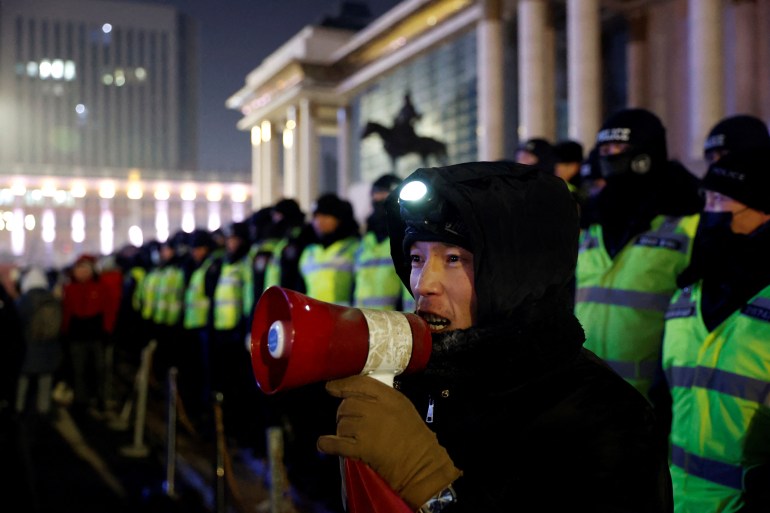 A protester in Mongolia with a small megaphone stands in front of a line of police officers in reflective vests.