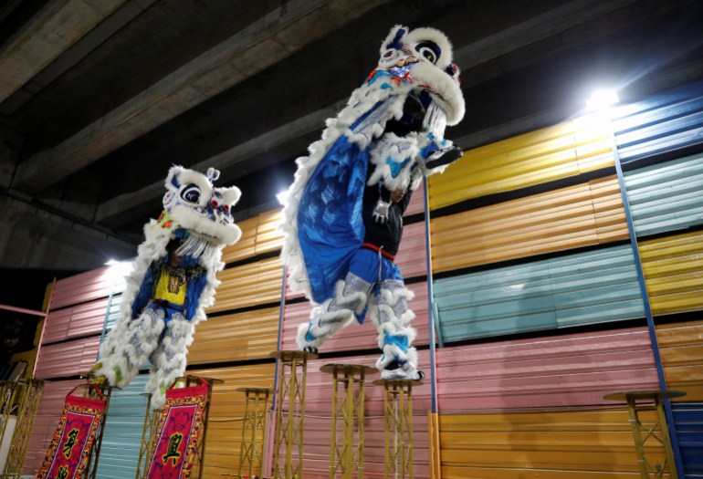 Two members of a lion dance troupe practice their routine ahead of the Lunar New Year.  They are wearing a bright blue costume fringed with white fur and each holding up the lion's head, which has large eyes with white fur around.  They are leaping across high poles