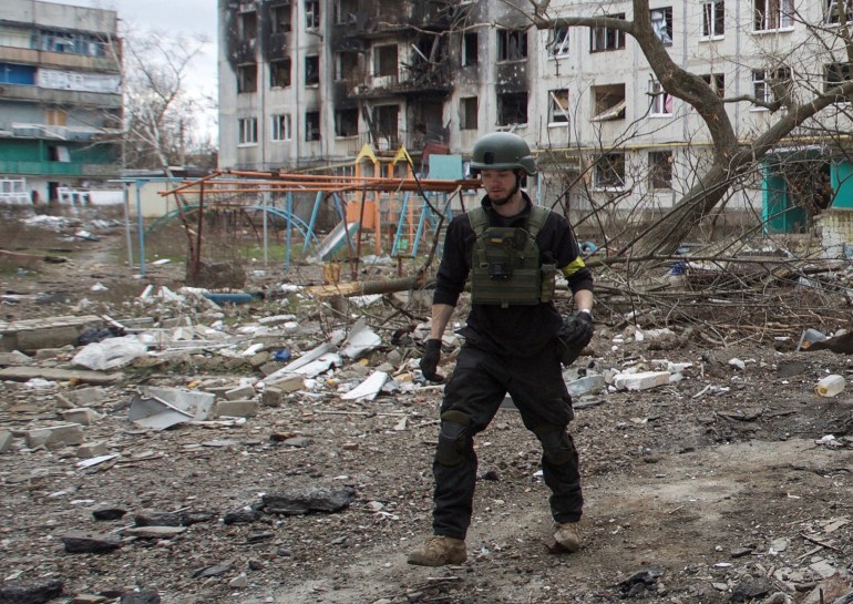 Christopher Parry in helmet and flak jacket walking outside a bombed out block of flats in Bakhmut. There is a mangled climbing frame behind him and the building is black where a missile hit