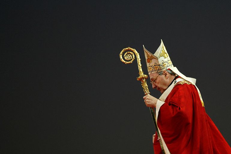 Cardinal George Pell in red robes as head of the Catholic Church in Australia. He is pictured in profile against a black backdrop and is carrying a gold staff in his left hand.
