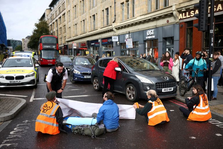 Protesters in orange high-vis jackets sit on a London street while a police officer is speaking to them, There are a number cars and a bus that have stopped.