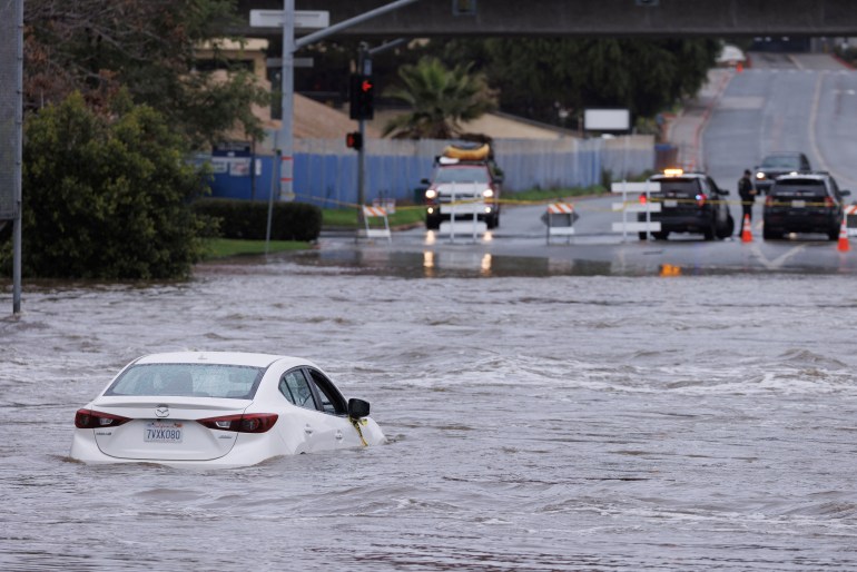 A white car sits in floodwater that reaches halfway up its doors in San Diego, California.  Emergency vehicles block the submerged road for a high point in the distance.