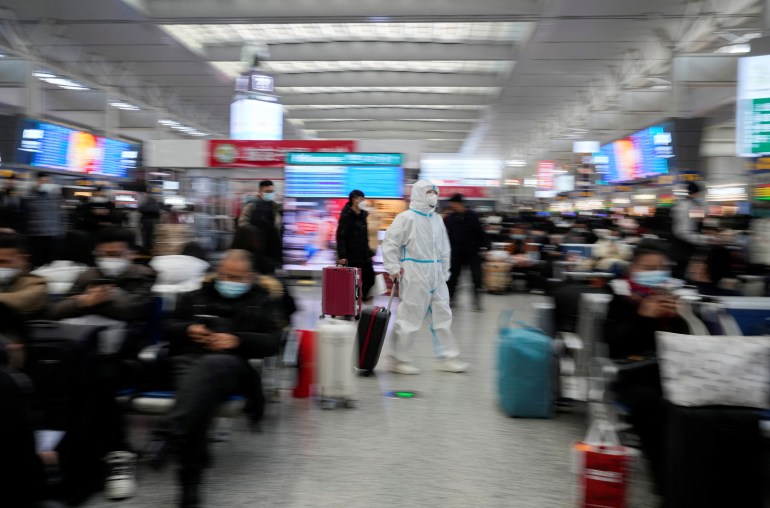 A woman at a Shanghai railway station walks with her luggage. There are lots of other passengers sitting with their suitcases. They are all wearing masks. The woman is in a white hazmat suit as well