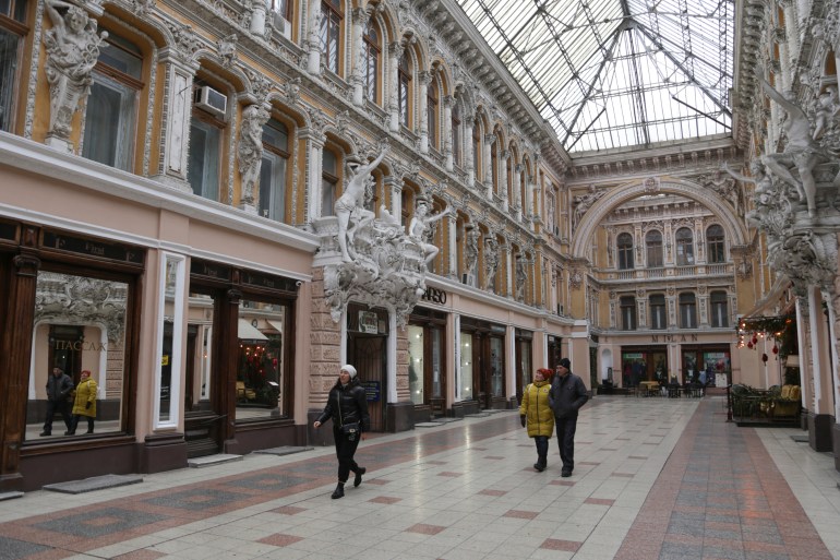 People walking in a glass-filled shopping arcade in the historic center of Odesa.  The buildings on either side are decorated and covered with statues.