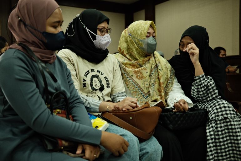 The four mothers sitting in court at a hearing for a class action suit they have brought against the government. They are wearing face masks and headscarves, and one is wearing a yellow t-shirt with a drawing of her son and the words 'my son is my hero'