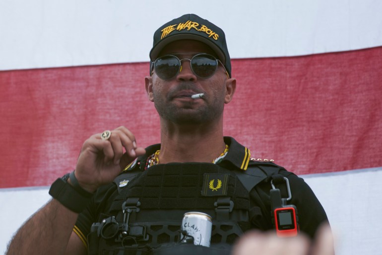 Enrique Tarrio, in a baseball cap and bulletproof vest. The black cap has yellow letters saying "THE WAR BOYS'. He is wearing sunglasses and has a cigarette in his mouth. There is a can of beer resting in a cup holder attached to his vest.