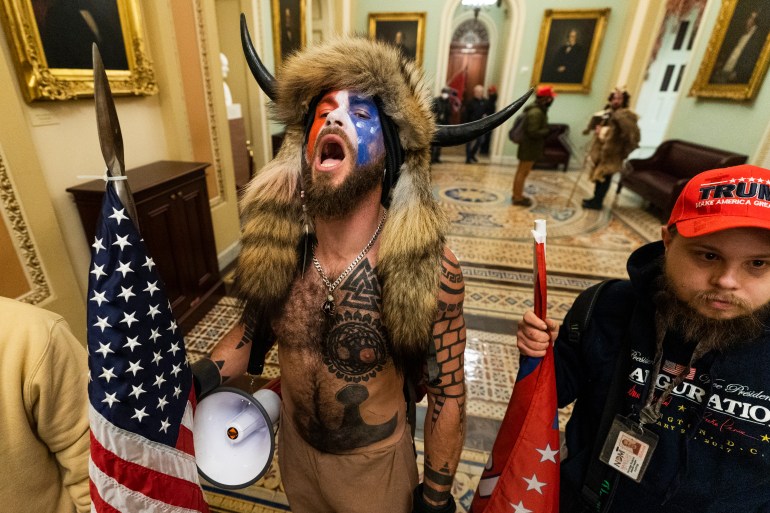 Jacob Chansley, with his top off and face painted with the US flag is looking upwards with his mouth open. He is wearing a horned helmet and fur hat.