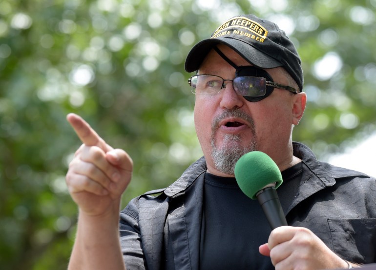 Stewart Rhodes, speaking into a microphone and pointing. He's wearing a black cap with Oath Keepers written across the front in yellow. He also has glasses on, which cover an eyepatch over his left eye.