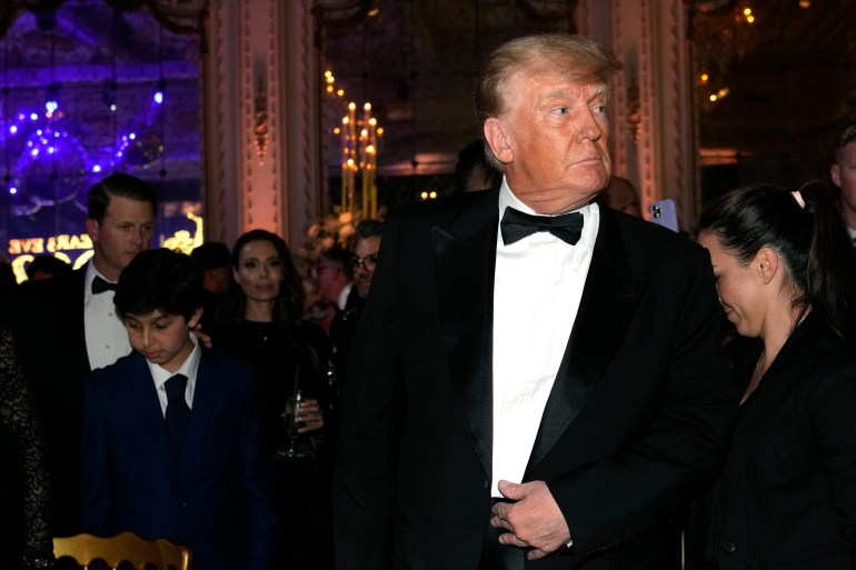 Trump wearing a tuxedo looks left. There are people behind him, one holding a champagne glass