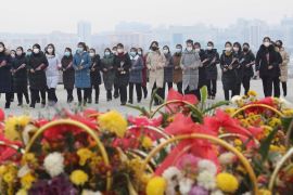 A group of North Koreans pictured behind a pile of bouquets laid on the ground during Lunar New Year. They are all wearing face masks. The outline of city buildings can be seen behind them through the haze.