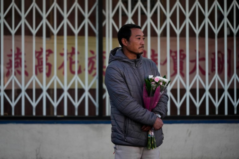 A man stands with a bouquet of white flowers outside a closed shop. He looks sad.