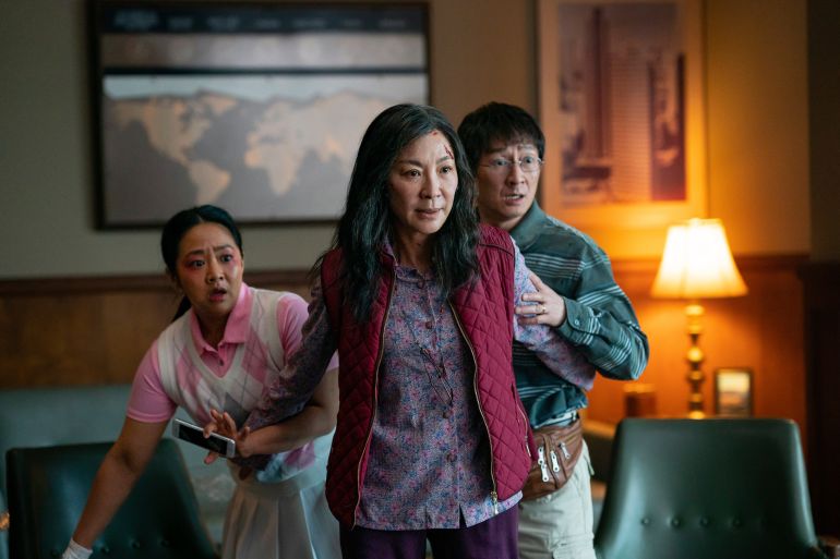 A still from Everything Everywhere All At Once showing Stephanie Hsu, Michelle Yeoh and Ke Huy Quan. They are in a living room. Yeoh's character appears to be protecting the others who are standing behind her looking worried
