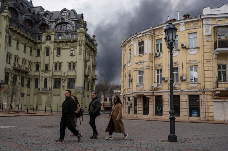 Historic buildings in the centre of Odesa, Ukraine. People are walking on a cobbled street. There is black smoke rising in the distance behind them.