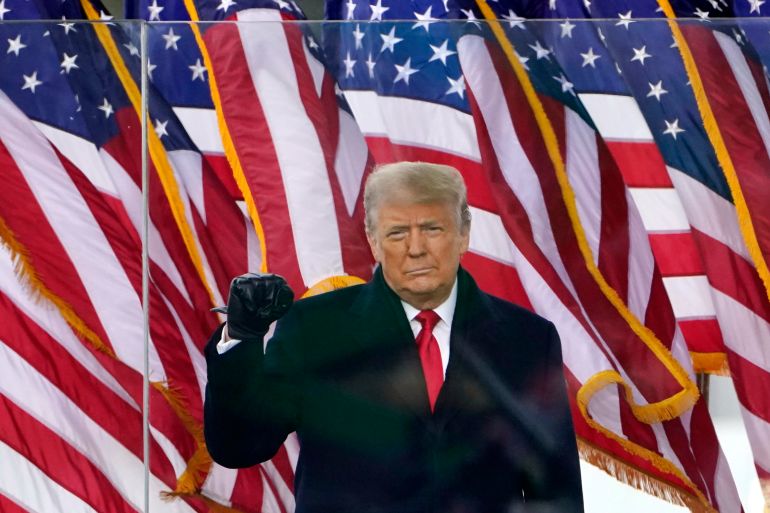 Donald Trump standing in front of some US flags. He's wearing a black coat and leather gloves. He is clenching his fist and looking serious