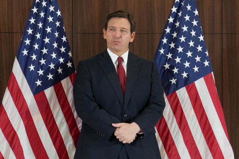 Florida Governor Ron DeSantis standing with hands folded between two US flags