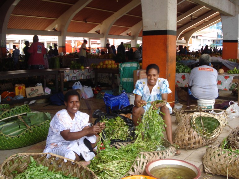 Two women in Vanuatu look at the camera while working in a covered market.
