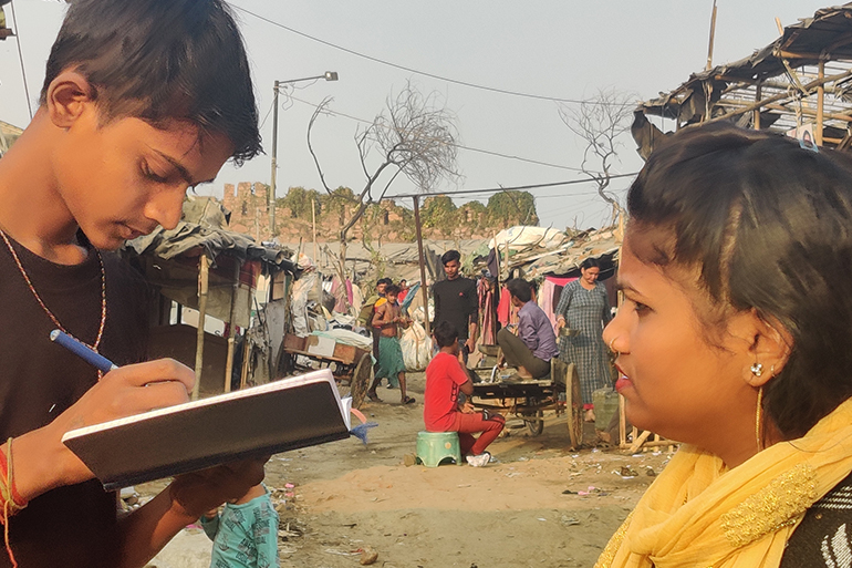 A photo of a boy writing something in a notebook with a woman standing in front of him talking.