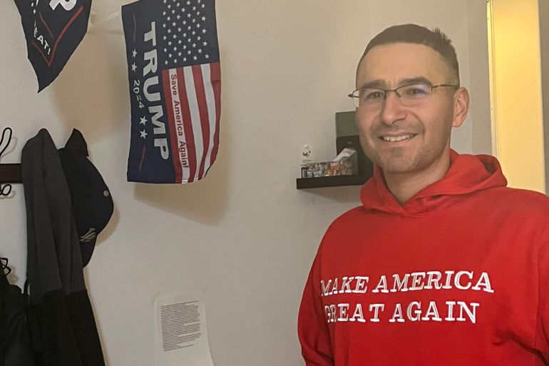 Solomon Pena wearing a red 'Make America Great Again' hoodie and a medium-sized Trump campaign flag hanging on the wall behind him.