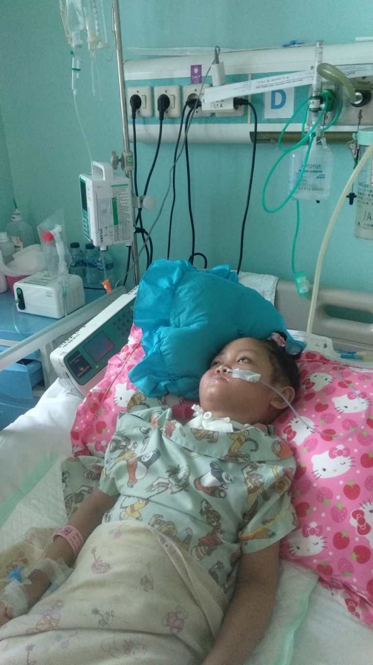 Shena lying in her hospital bed. She has a tube to her nose and a cannula in her hand. She is lying on her back with head supported to the right by a pillow. Her eyes are open but she is looking up at the ceiling