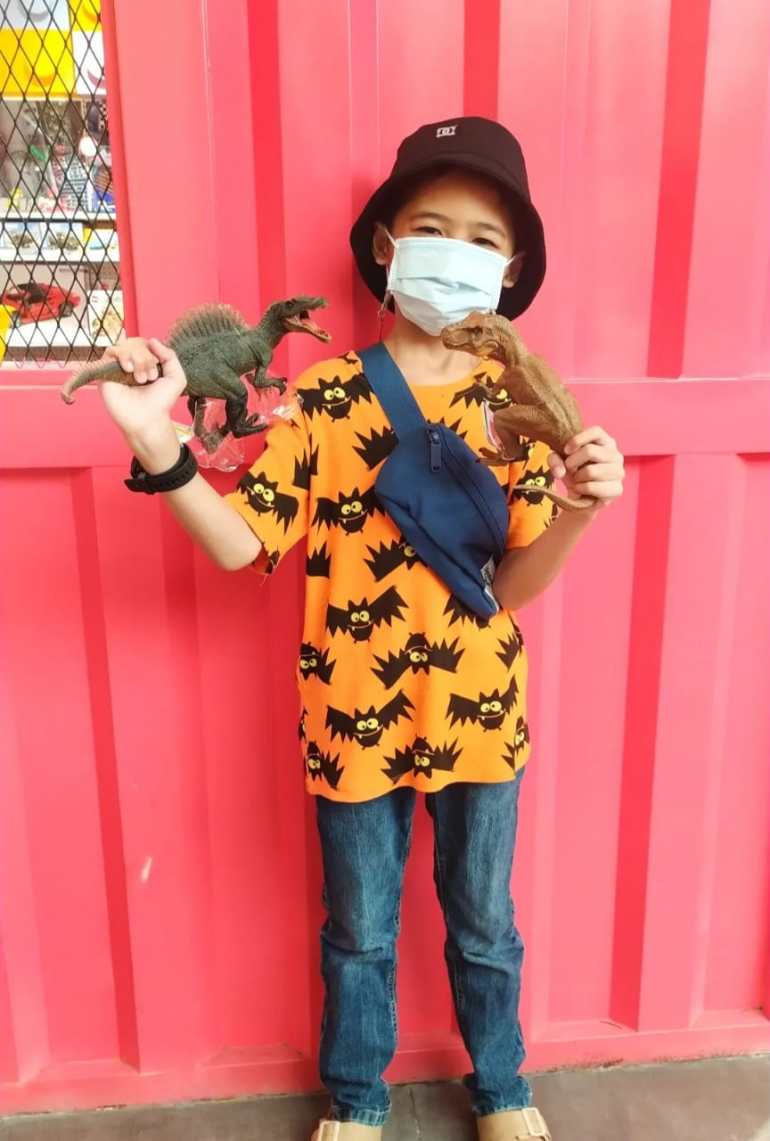 Panghegar standing in front of the red wall.  He was wearing an orange t-shirt with a print of black cartoon bats and jeans.  He has a hat on his head and holds a toy dinosaur in each of his hands.