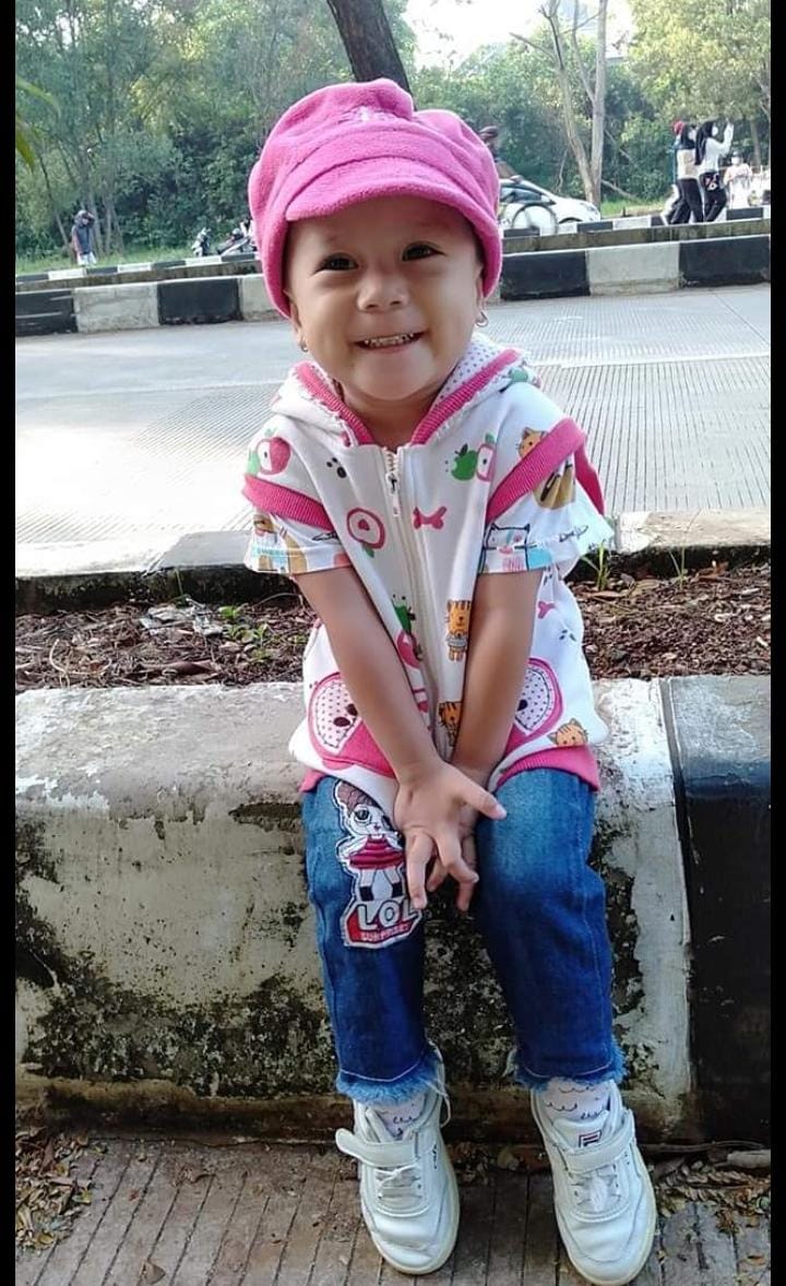 Azqiara sitting on the kerb by the side of the road.  She is wearing jeans, a pink and white short and a pink hat.  She is smiling and looks really happy