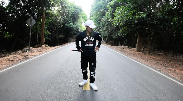 Kea Sokun in black sweatpants and sweatshirt with the word WONDER written on the chest.  He performs in a rap video.  He is standing on a road with trees on either side.  He has his hands on his hips and holds a microphone in his right hand and looks down and away from the camera on his left.  He wears a white baseball cap.  His trainers are also white.