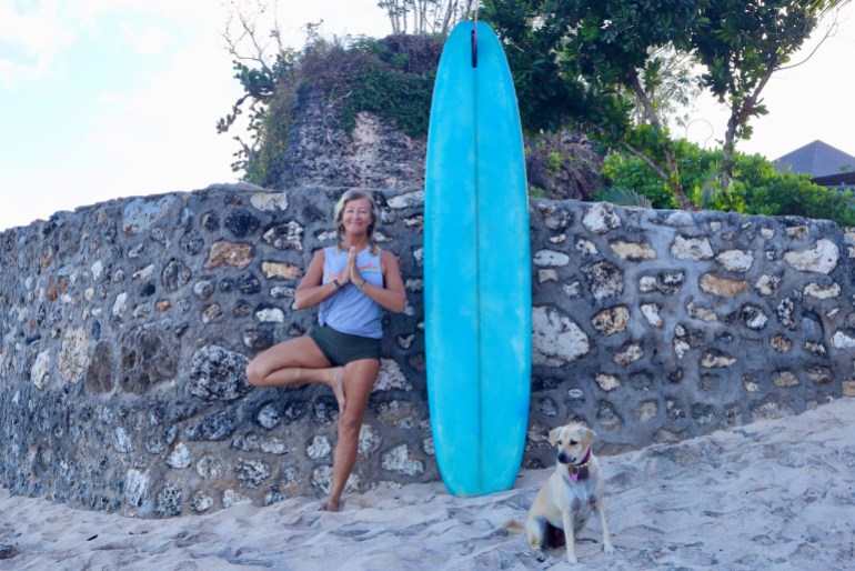 Kit Cahill leans against a rock wall in a yoga pose with one foot in the sand with a surfboard standing beside her and a medium-sized dog looking into the distance.